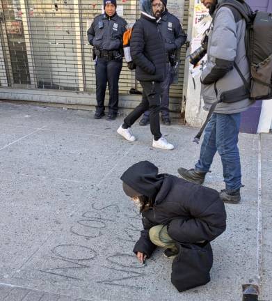 A masked and hooded female protestor writes NO COPS NO KKK in chalk on the sidewalk. Behind her stand two uniformed NYPD officers, one an African-American woman, the other an Indian-American man.